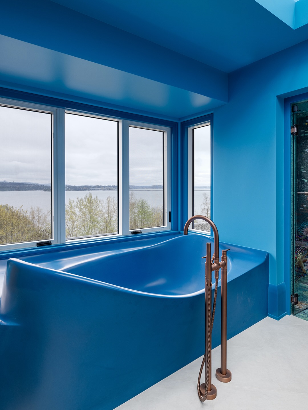 Bespoke blue freestanding cast bathtub with copper floor-mount faucet, Puget Sound visible through the window